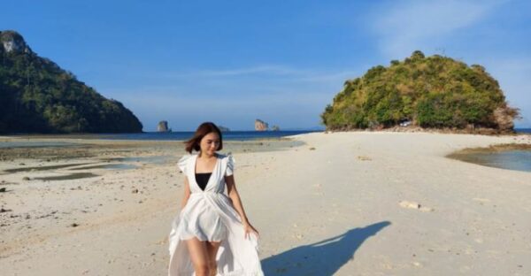 A woman in a white dress walks along the beach, enjoying the serene atmosphere and the sound of the ocean.