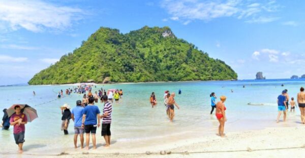 A crowded beach scene with numerous people enjoying the sun and sand near a picturesque island in the background.