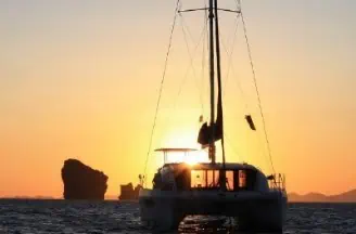 A catamaran sailing at sunset with rocky formations in the background and a golden sky reflecting on calm waters in Phuket.