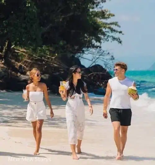 Three individuals stroll along the beach, each holding a drink, enjoying a sunny day by the ocean.