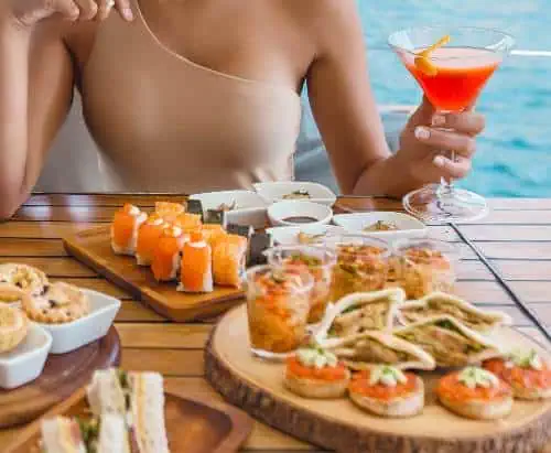 A woman sits at a table filled with food, holding a cocktail and savoring the moment.