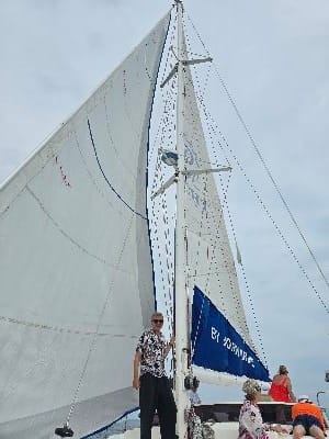 Young man in front of the white sails of a catamaran, with the vast sea and clear sky in the background.