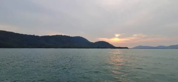 Thailändisches Boot auf stillem Meer bei Sonnenuntergang, Berge im Hintergrund in goldenem Licht.