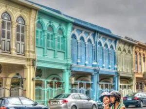 A happy couple riding a motorbike in front of vibrant, colorful buildings, showcasing the spirit of adventure in Phuket.