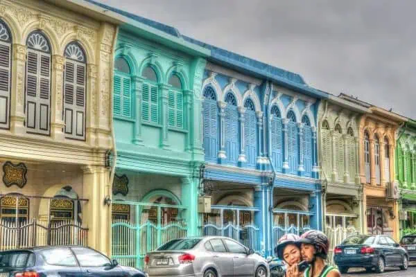 A happy couple riding a motorbike in front of vibrant, colorful buildings, showcasing the spirit of adventure in Phuket.