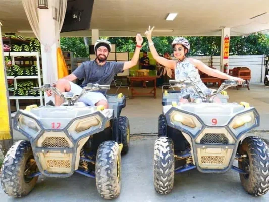 A man and woman riding ATVs in a parking lot, surrounded by parked vehicles and clear blue skies.