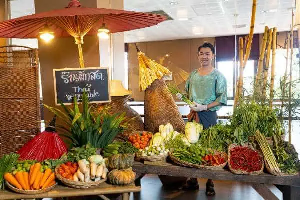 A woman stands before a vibrant vegetable display at the Siam Niramit Show in Phuket, showcasing local produce.