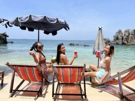 Three women enjoying drinks while sitting on the beach, surrounded by sand and ocean waves under a clear sky.
