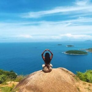 A woman sits atop a mountain, gazing out over the vast ocean, surrounded by breathtaking natural beauty.